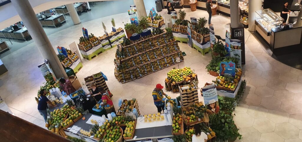 Puestos de stand de diferente tamaño de fruta exotica como piña, mango y papaya en una zona de entrada de un supermercado. Además se ve una zona donde se corta y vende la fruta recién cortada